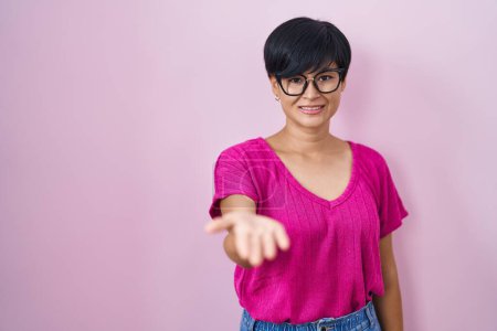 Foto de Joven mujer asiática con pelo corto de pie sobre fondo rosa sonriente alegre ofreciendo la palma de la mano dando asistencia y aceptación. - Imagen libre de derechos