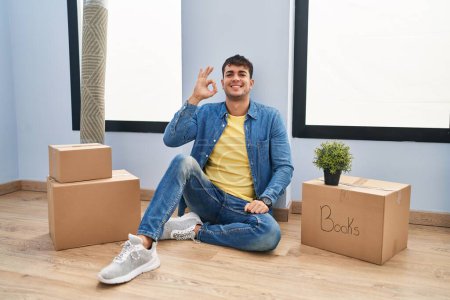 Photo for Young hispanic man sitting on the floor at new home smiling positive doing ok sign with hand and fingers. successful expression. - Royalty Free Image