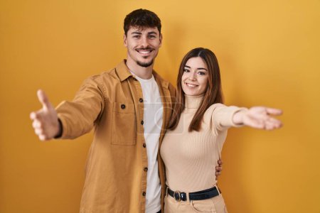 Téléchargez les photos : Jeune couple hispanique debout sur fond jaune regardant la caméra souriant à bras ouverts pour un câlin. expression joyeuse embrassant le bonheur. - en image libre de droit