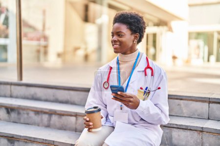 Photo for African american woman wearing doctor uniform using smartphone drinking coffee at street - Royalty Free Image