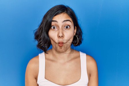 Photo for Young hispanic woman standing over blue background making fish face with lips, crazy and comical gesture. funny expression. - Royalty Free Image