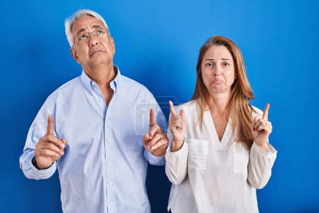 Téléchargez les photos : Couple hispanique du moyen âge debout sur fond bleu pointant vers le haut regardant triste et contrarié, indiquant la direction avec les doigts, malheureux et déprimé. - en image libre de droit