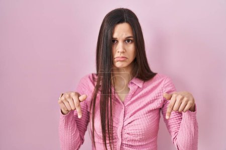 Foto de Young hispanic woman standing over pink background pointing down looking sad and upset, indicating direction with fingers, unhappy and depressed. - Imagen libre de derechos
