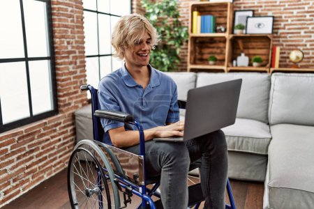 Photo for Young blond man using laptop sitting on wheelchair at home - Royalty Free Image