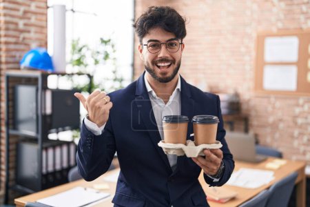 Foto de Hombre hispano con barba trabajando en la oficina sosteniendo el café apuntando con el pulgar hacia un lado sonriendo feliz con la boca abierta - Imagen libre de derechos