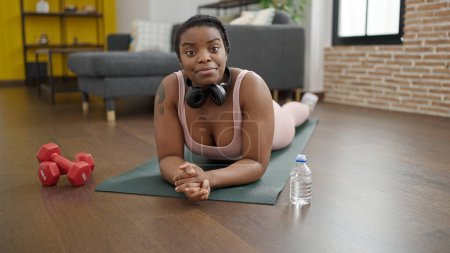 Téléchargez les photos : Femme afro-américaine souriante confiante allongée sur un tapis de yoga à la maison - en image libre de droit