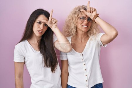 Téléchargez les photos : Mère et fille debout ensemble sur fond rose se moquant des gens avec les doigts sur le front faisant geste de perdant se moquant et insultant. - en image libre de droit