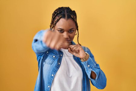 Téléchargez les photos : Femme afro-américaine avec des tresses debout sur fond jaune coups de poing poing pour se battre, agressif et en colère attaque, menace et violence - en image libre de droit