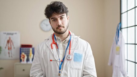 Foto de Joven médico hispano de pie con expresión seria en la clínica - Imagen libre de derechos