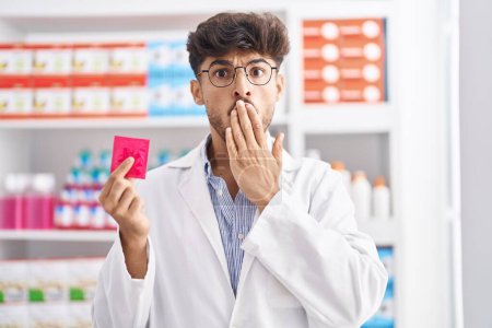 Téléchargez les photos : Arabe homme avec barbe travaillant à la pharmacie pharmacie tenant préservatif regardant la caméra souffler un baiser étant belle et sexy. expression d'amour. - en image libre de droit