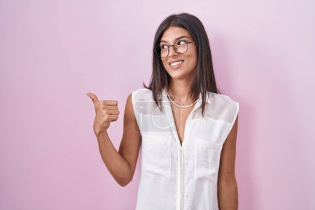 Photo for Brunette young woman standing over pink background wearing glasses smiling with happy face looking and pointing to the side with thumb up. - Royalty Free Image