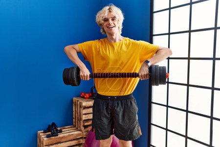 Téléchargez les photos : Jeune homme blond souriant confiant en utilisant la musculation au centre sportif - en image libre de droit