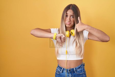 Foto de Mujer rubia joven de pie sobre fondo amarillo con auriculares haciendo pulgares hacia arriba y hacia abajo, el desacuerdo y la expresión de acuerdo. conflicto loco - Imagen libre de derechos