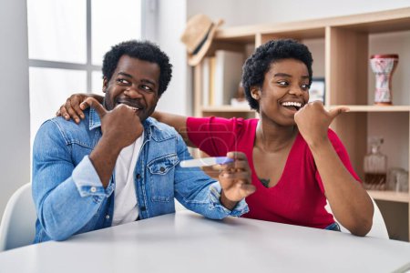 Téléchargez les photos : Jeune couple afro-américain tenant le résultat du test de grossesse pointant le pouce vers le côté souriant heureux avec la bouche ouverte - en image libre de droit