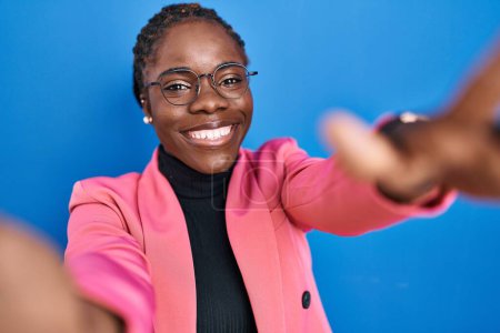 Téléchargez les photos : Beautiful black woman standing over blue background looking at the camera smiling with open arms for hug. cheerful expression embracing happiness. - en image libre de droit