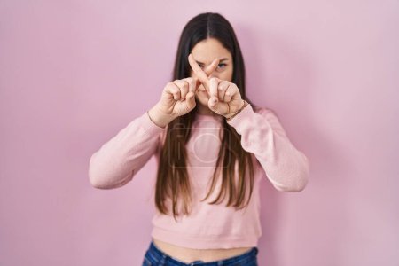 Foto de Joven morena de pie sobre fondo rosa rechazo expresión cruzando dedos haciendo signo negativo - Imagen libre de derechos