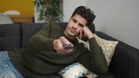 Photo for Young hispanic man watching tv lying on sofa with boring expression at home - Royalty Free Image