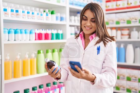 Téléchargez les photos : Jeune belle pharmacienne hispanique tenant un flacon de médicaments à l'aide d'un smartphone à la pharmacie - en image libre de droit