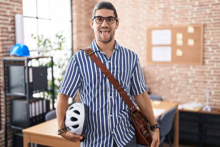 Photo for Hispanic man with long hair working at the office holding bike helmet sticking tongue out happy with funny expression. emotion concept. - Royalty Free Image