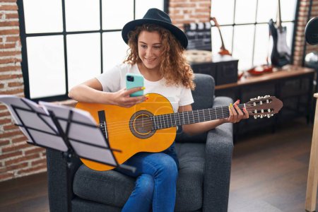 Photo for Young beautiful hispanic woman musician playing classical guitar using smartphone at music studio - Royalty Free Image