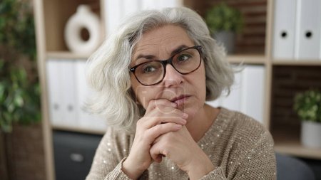Téléchargez les photos : Femme d'âge moyen avec des cheveux gris travailleur d'affaires assis avec l'expression du doute au bureau - en image libre de droit