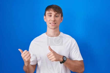 Photo for Caucasian blond man standing over blue background pointing to the back behind with hand and thumbs up, smiling confident - Royalty Free Image