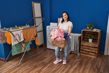 Foto de Síndrome de Down mujer sonriendo con confianza celebración cesta con ropa en la sala de lavandería - Imagen libre de derechos