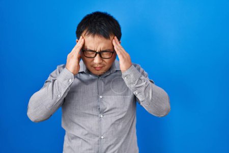 Foto de Joven hombre chino de pie sobre fondo azul con la mano en la cabeza para el dolor en la cabeza porque el estrés. padecer migraña. - Imagen libre de derechos