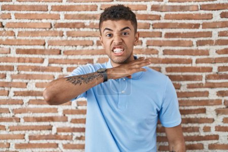 Photo for Brazilian young man standing over brick wall cutting throat with hand as knife, threaten aggression with furious violence - Royalty Free Image