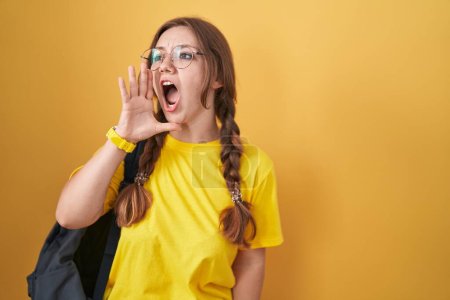 Photo for Young caucasian woman wearing student backpack over yellow background shouting and screaming loud to side with hand on mouth. communication concept. - Royalty Free Image