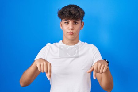 Photo for Hispanic teenager standing over blue background pointing down looking sad and upset, indicating direction with fingers, unhappy and depressed. - Royalty Free Image