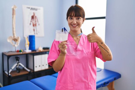 Photo for Young brunette woman working at rehabilitation clinic showing id smiling happy and positive, thumb up doing excellent and approval sign - Royalty Free Image