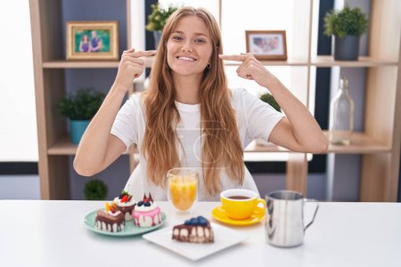 Téléchargez les photos : Jeune femme caucasienne manger des pâtisseries t pour le petit déjeuner sourire gai montrant et pointant avec les doigts dents et la bouche. concept de santé dentaire. - en image libre de droit