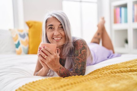Téléchargez les photos : Femme aux cheveux gris du moyen âge buvant une tasse de café allongée sur le lit dans la chambre - en image libre de droit