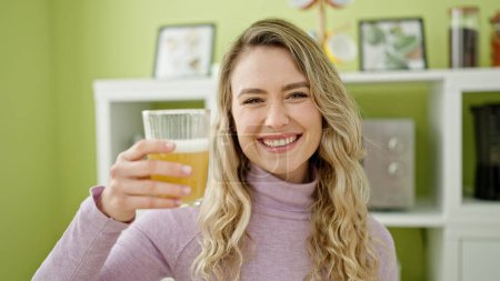 Photo for Young blonde woman holding glass of orange juice sitting on table at dinning room - Royalty Free Image