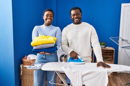 Téléchargez les photos : Jeune couple afro-américain repassage vêtements à la buanderie sourire avec un sourire heureux et cool sur le visage. montrant les dents. - en image libre de droit
