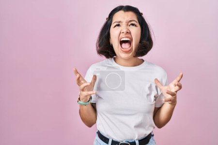 Foto de Mujer hispana joven con camiseta blanca casual sobre fondo rosa loca y loca gritando y gritando con expresión agresiva y los brazos levantados. concepto de frustración. - Imagen libre de derechos