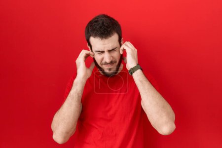 Foto de Joven hombre hispano con camiseta roja casual cubriendo las orejas con los dedos con expresión molesta por el ruido de la música alta. concepto de sordo. - Imagen libre de derechos