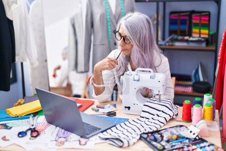 Foto de Middle age grey-haired woman tailor using sewing machine and laptop at tailor shop - Imagen libre de derechos