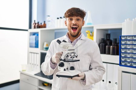 Téléchargez les photos : Homme arabe avec barbe travaillant au laboratoire scientifique tenant le microscope célébrant fou et étonné pour le succès avec les yeux ouverts hurlant excité. - en image libre de droit