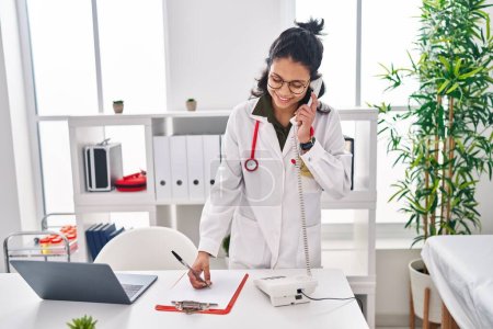 Téléchargez les photos : Jeune femme latine médecin parlant au téléphone écrit sur le document à la clinique - en image libre de droit