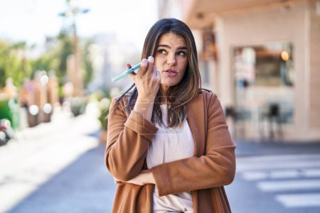 Téléchargez les photos : Jeune belle femme hispanique parlant sur smartphone avec une expression sérieuse dans la rue - en image libre de droit