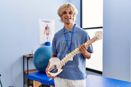 Foto de Joven pisioterapeuta hombre rubio sonriendo confiado sosteniendo modelo anatómico de columna vertebral en clínica de rehabilitación - Imagen libre de derechos