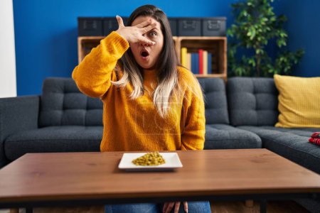 Foto de Mujer hispana joven comiendo guisantes verdes asomándose en shock cubriendo la cara y los ojos con la mano, mirando a través de los dedos asustados - Imagen libre de derechos