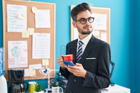 Photo for Young hispanic man business worker using smartphone drinking coffee at office - Royalty Free Image