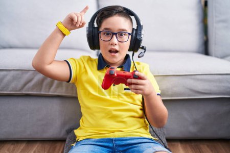Foto de Joven niño hispano jugando videojuego sosteniendo controlador usando auriculares sonriendo asombrado y sorprendido y señalando con los dedos y los brazos levantados. - Imagen libre de derechos