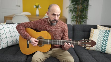 Photo for Young bald man playing classical guitar sitting on sofa at home - Royalty Free Image