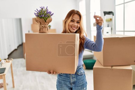 Foto de Joven mujer caucásica sonriendo confiado sosteniendo paquete con planta de lavanda y llave en el nuevo hogar - Imagen libre de derechos