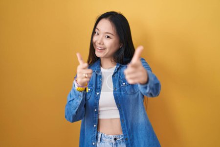 Photo for Young asian woman standing over yellow background pointing fingers to camera with happy and funny face. good energy and vibes. - Royalty Free Image