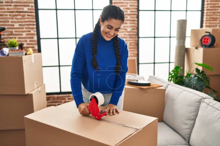 Foto de Young hispanic woman smiling confident packaging cardboard box at new home - Imagen libre de derechos